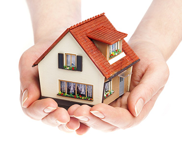 A woman holding a model house in her hands, representing the services provided by Ideal Insurance Agency to protect homeowners.