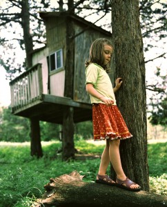 A little girl standing on a log in front of a tree house.