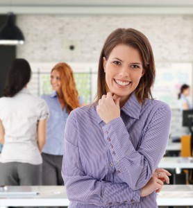 A smiling business woman standing in an office with her arms crossed.