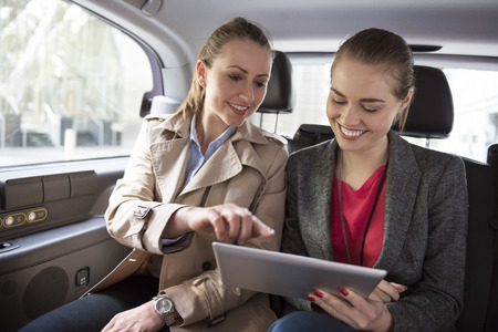 Two women sitting in the back seat of a car looking at a tablet.