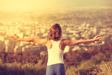 A woman with her arms outstretched on top of a hill overlooking a city.